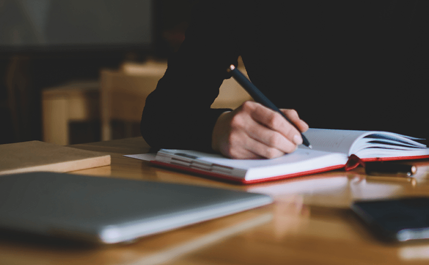 Close-up of a hand writing in a notebook on a wooden desk.
