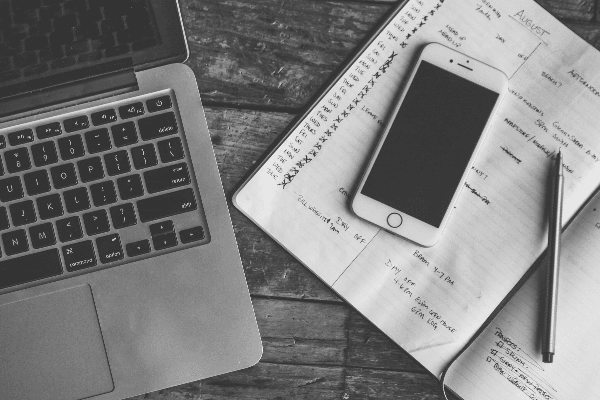 Overhead black-and-white photo of a laptop, smartphone, and notebook on a wooden desk.