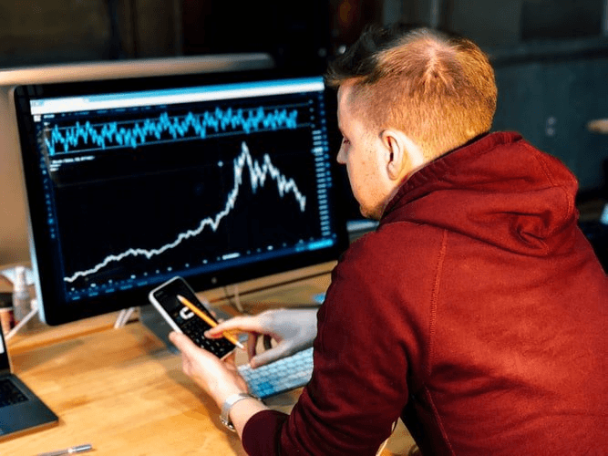 Trader reviewing a price chart on a desktop monitor while holding a smartphone.