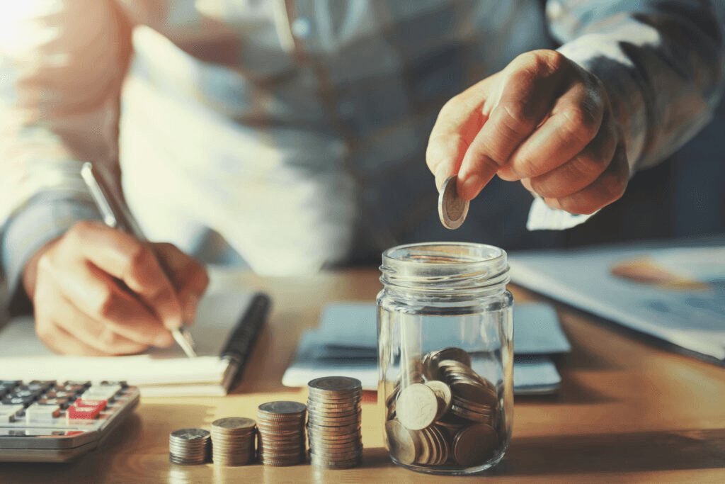 Person placing coins into a glass jar on a desk with a calculator and paperwork.