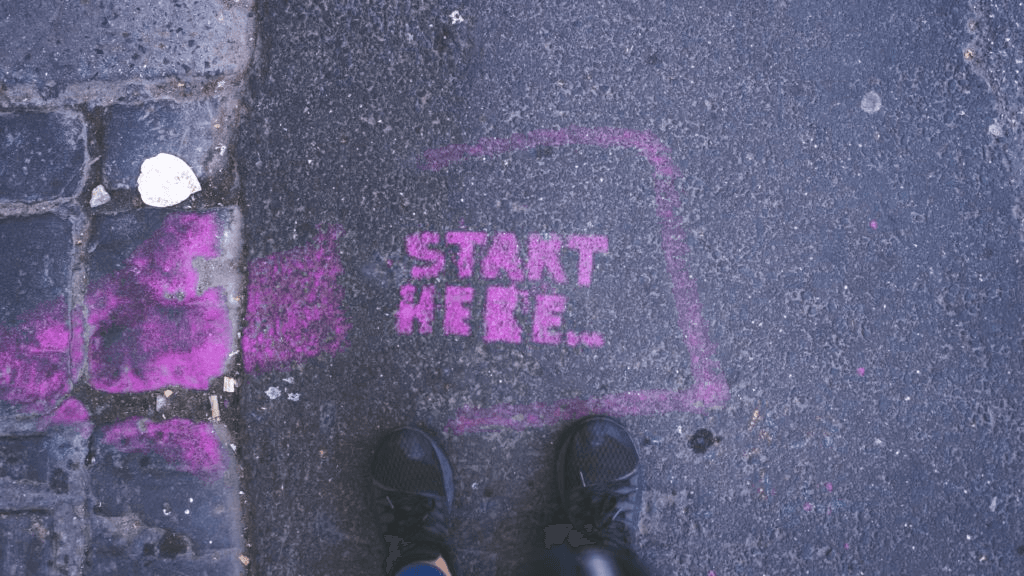 Chalk text reading “Start Here” on pavement with shoes visible.